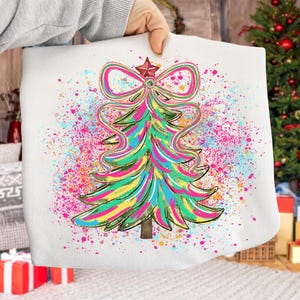 May include: White Christmas-themed tote bag featuring a colorful Christmas tree design. The tree is decorated with a bow and star, and surrounded by a vibrant splatter of pink, blue, and yellow paint. The bag is held by a person.