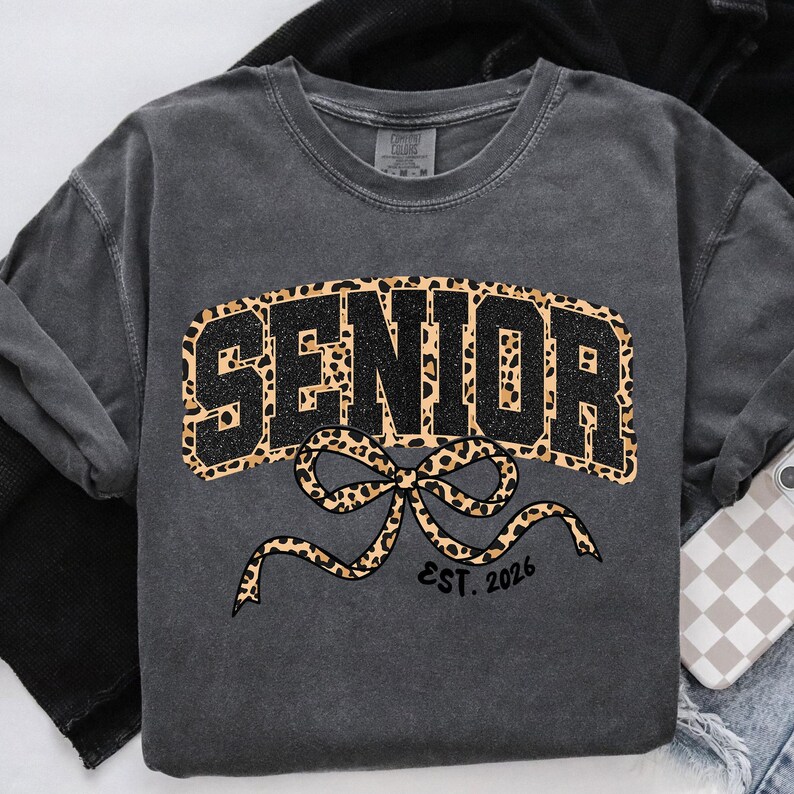 May include: Dark gray sweatshirt with the word "SENIOR" in black glitter and leopard print. A leopard print bow is below the word, with "EST. 2026" printed below the bow. The sweatshirt is a crew neck style.