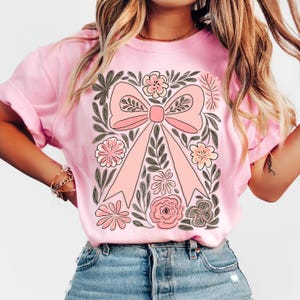 K&ouml;nnte beinhalten: Hellrosa T-Shirt mit einem gro&szlig;en, zentralen rosa Schleifendesign, umgeben von floralen und Blattmotiven in Rosa, Grau und Wei&szlig;. Das T-Shirt hat kurze &Auml;rmel und einen Rundhalsausschnitt. Das Thema ist floral und feminin.