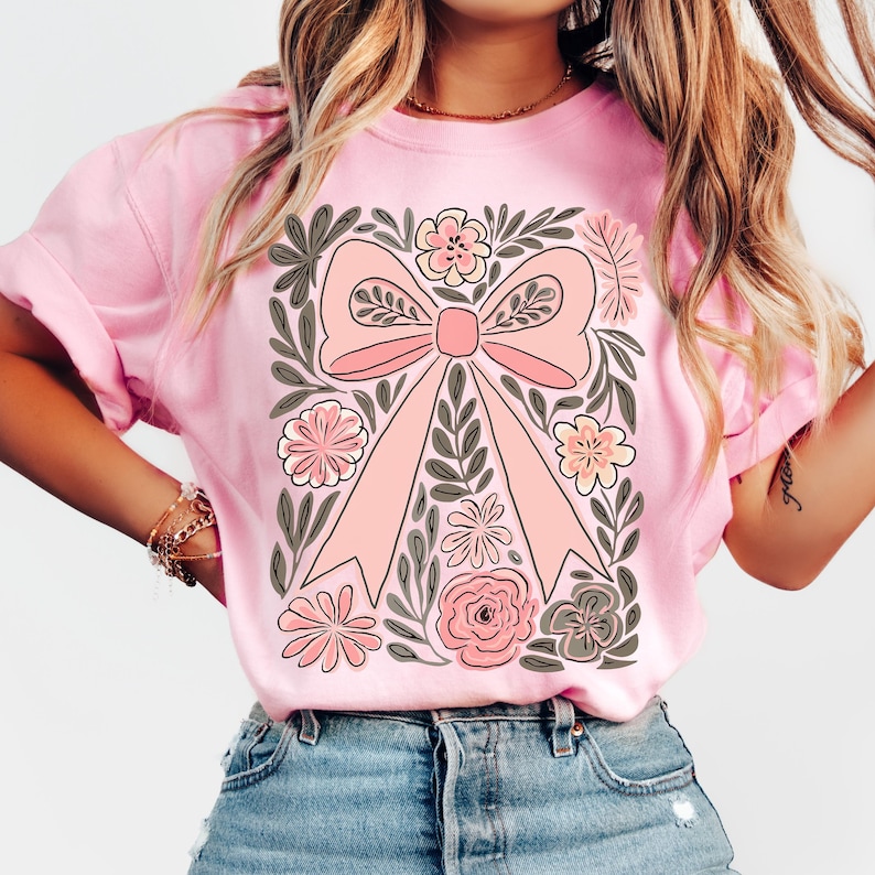 K&ouml;nnte beinhalten: Hellrosa T-Shirt mit einem gro&szlig;en, zentralen rosa Schleifendesign, umgeben von floralen und Blattmotiven in Rosa, Grau und Wei&szlig;. Das T-Shirt hat kurze &Auml;rmel und einen Rundhalsausschnitt. Das Thema ist floral und feminin.