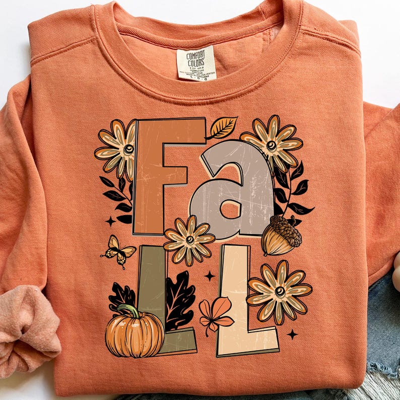 May include: An orange Comfort Colors sweatshirt with a fall-themed graphic. The graphic includes the word "FALL" in brown and tan letters, surrounded by flowers, leaves, a pumpkin, an acorn, and a butterfly.