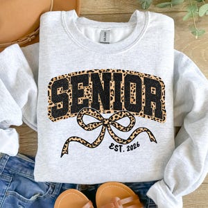 May include: A light gray sweatshirt with the word "SENIOR" in black glitter letters outlined with a leopard print design. A leopard print bow with the text "EST. 2026" is below the word "SENIOR".