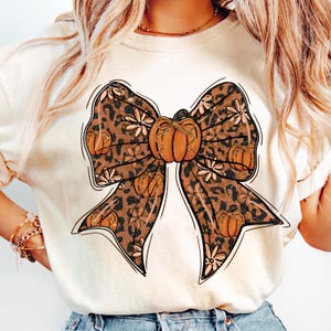 May include: Cream-colored t-shirt featuring a large bow design with a pumpkin in the center. The bow has a leopard print pattern with pumpkins and floral accents. The design is outlined in black.