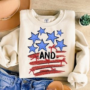 Pode incluir: Uma camisa de moletom cor creme com um design de bandeira americana vermelha, branca e azul. A bandeira &eacute; feita de listras vermelhas e brancas com estrelas azuis. A palavra "AND" est&aacute; escrita em letras pretas na bandeira.