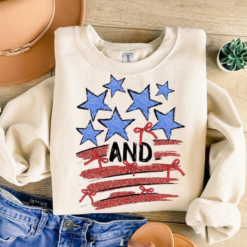 Pode incluir: Uma camisa de moletom cor creme com um design de bandeira americana vermelha, branca e azul. A bandeira &eacute; feita de listras vermelhas e brancas com estrelas azuis. A palavra "AND" est&aacute; escrita em letras pretas na bandeira.