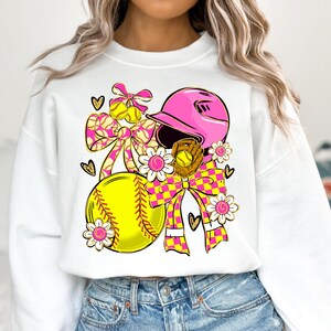 May include: White sweatshirt featuring a softball design. The design includes a pink helmet, a yellow softball, a glove, and decorative bows with a yellow and pink checkered pattern. Floral and heart accents complete the design.