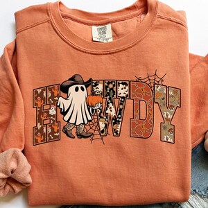 Puede incluir: Sudadera naranja Comfort Colors con un gr&aacute;fico tem&aacute;tico de Halloween. El gr&aacute;fico presenta un fantasma con sombrero y botas de vaquero, sosteniendo una calabaza, con la palabra "HOWDY" en una fuente decorativa. Tambi&eacute;n se incluye una telara&ntilde;a.