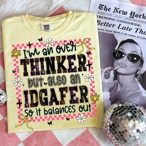 May include: A light yellow t-shirt with the text "I'm an over thinker but also an IDGAFER so it balances out" in black and pink. The shirt has a crew neck and short sleeves. The design includes flowers, butterflies, and bows.