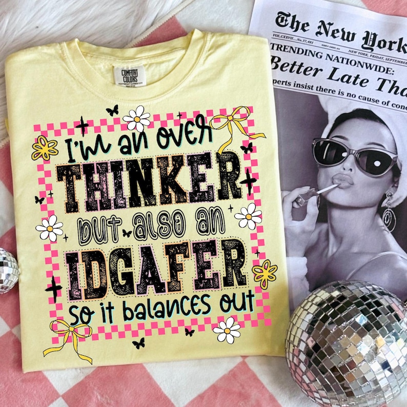 May include: A light yellow t-shirt with the text "I'm an over thinker but also an IDGAFER so it balances out" in black and pink. The shirt has a crew neck and short sleeves. The design includes flowers, butterflies, and bows.