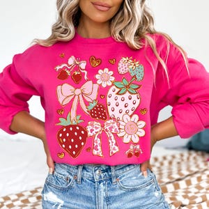 Op de afbeelding: Een roze sweatshirt met een grafische print van aardbeien, strikken en bloemen. Het ontwerp is in tinten roze, rood, groen en goud.