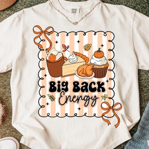 May include: Cream-colored t-shirt with a graphic featuring fall food illustrations and the text "Big Back Energy." The design includes a pumpkin spice latte, pie, roasted turkey, and a cupcake with a pumpkin on top. Orange bows and a striped background complete the design.