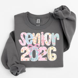 Puede incluir: Una sudadera gris oscuro con las palabras "Senior 2026" en un dise&ntilde;o de patchwork floral colorido. El texto incluye patrones florales rosas, azules y amarillos, con un lazo rosa que acent&uacute;a el "2026". La sudadera tiene cuello redondo y mangas largas.