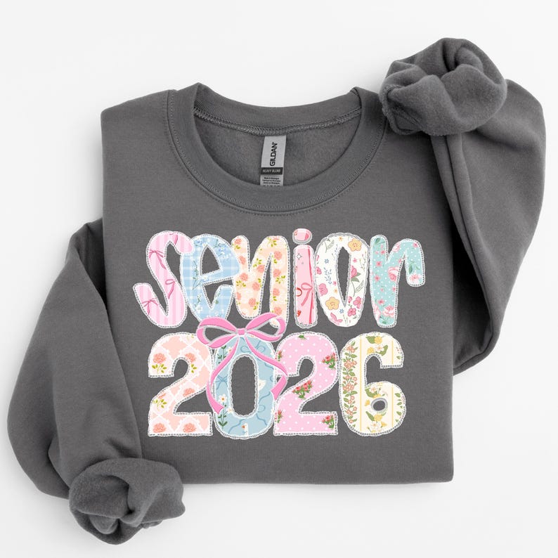 Puede incluir: Una sudadera gris oscuro con las palabras "Senior 2026" en un dise&ntilde;o de patchwork floral colorido. El texto incluye patrones florales rosas, azules y amarillos, con un lazo rosa que acent&uacute;a el "2026". La sudadera tiene cuello redondo y mangas largas.