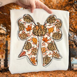 May include: A white sweatshirt featuring a large bow design with a pumpkin in the center. The bow is decorated with leopard print pumpkins, orange maple leaves, and floral patterns. The design has a fall or autumn theme.