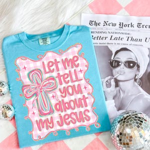May include: A light blue t-shirt with the text "Let me tell you about my Jesus" in pink and white. The design includes a cross with a bow and a gingham pattern. The shirt is on a pink and white checkered surface.
