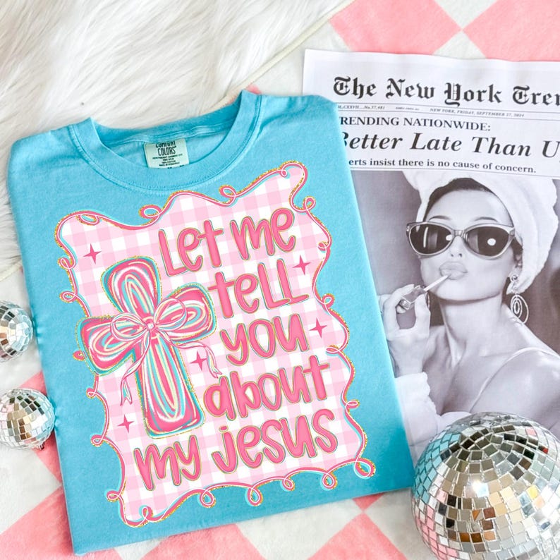 May include: A light blue t-shirt with the text "Let me tell you about my Jesus" in pink and white. The design includes a cross with a bow and a gingham pattern. The shirt is on a pink and white checkered surface.