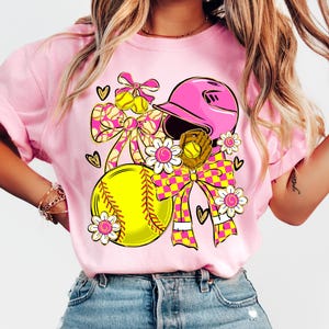 May include: Pink t-shirt featuring a softball design. The design includes a yellow softball, pink helmet, glove, and bows with a checkered pattern. White daisies and hearts complete the design.