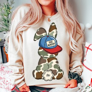 May include: A beige sweatshirt with a cartoon bunny wearing a blue and red baseball cap that says "Egg Hunter". The bunny is wearing camouflage.