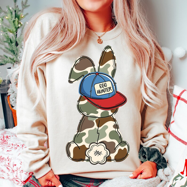 May include: A beige sweatshirt with a cartoon bunny wearing a blue and red baseball cap that says "Egg Hunter". The bunny is wearing camouflage.