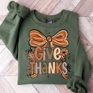 May include: An olive green sweatshirt with the words "Give Thanks" in a fall-themed design. The graphic includes a large bow, flowers, and leaves in shades of orange, brown, and beige. The sweatshirt is made of a soft material.