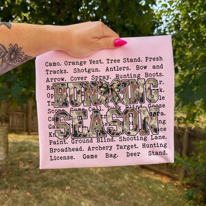 May include: A light pink t-shirt with the words "Hunting Season" in camouflage print. The shirt also lists hunting-related terms such as "Shotgun", "Antlers", and "Game Bag".