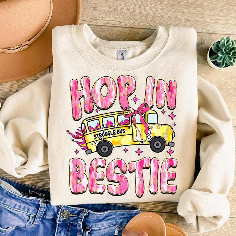 May include: A cream colored sweatshirt with a pink and yellow graphic design. The graphic features the text "Hop In Bestie" and a yellow school bus with the text "Struggle Bus" on the side.