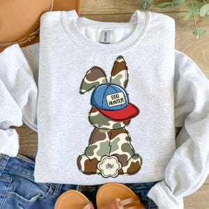 May include: A light gray sweatshirt with a cartoon image of a camouflage bunny wearing a blue and red baseball cap that says "Egg Hunter".