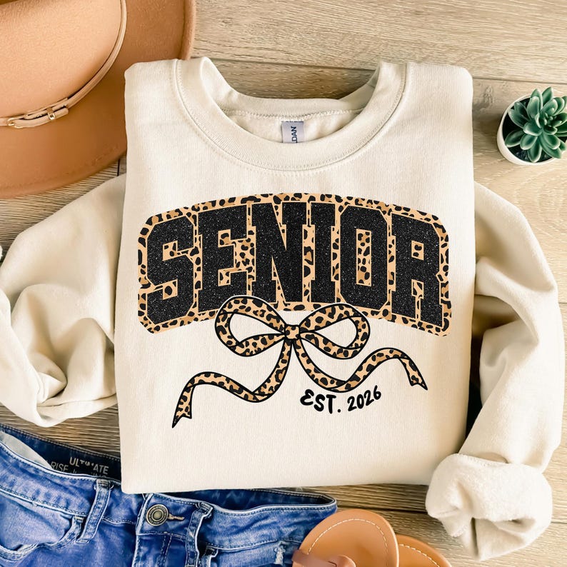 May include: Cream-colored sweatshirt with the word "SENIOR" in a black and leopard print design. A leopard print bow is below the word, with "EST. 2026" printed below the bow. The sweatshirt is laid flat on a wooden surface.