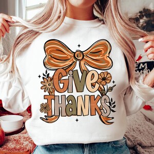 May include: White sweatshirt with a large graphic that says "Give Thanks" in various fall colors. The design includes a bow, flowers, and leaves. The sweatshirt is a long-sleeved top.