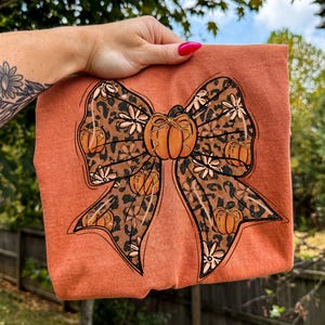 May include: Rust-colored t-shirt featuring a large bow design with a pumpkin in the center. The bow has a leopard print pattern with small pumpkins and white flowers. The shirt is held up by a hand with pink nail polish.