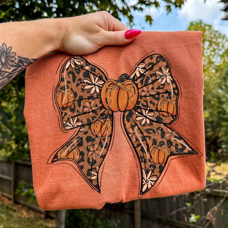 May include: Rust-colored t-shirt featuring a large bow design with a pumpkin in the center. The bow has a leopard print pattern with small pumpkins and white flowers. The shirt is held up by a hand with pink nail polish.