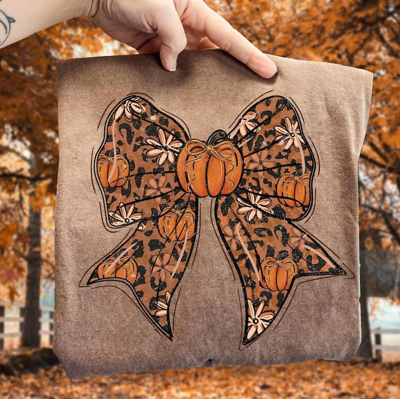 May include: A brown sweatshirt featuring a large bow design with a leopard print pattern. The bow is adorned with orange pumpkins and white floral accents, perfect for fall. The design is held up by a hand.
