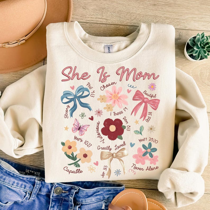 K&ouml;nnte beinhalten: Ein cremefarbener Pullover mit dem Text "She Is Mom" in rosa Schrift. Der Pullover ist mit Blumenmustern, Schmetterlingen, Schleifen und Bibelversen verziert.