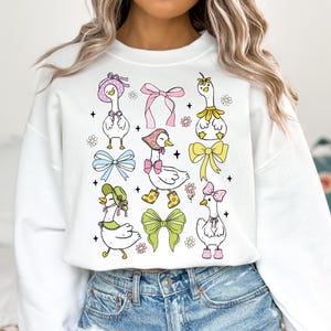 Puede incluir: Sudadera blanca con un estampado gr&aacute;fico colorido de gansos con sombreros y lazos. Los gansos est&aacute;n rodeados de flores y estrellas.