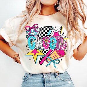Op de afbeelding: Cr&egrave;mekleurig T-shirt met een levendige "Cheer"-afbeelding. Het ontwerp bevat een ster, megafoon en strik in roze, blauw, geel en zwart. Het woord "Cheer" is in een vet, kleurrijk lettertype, perfect voor cheerleading.