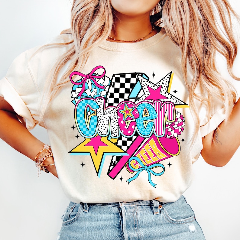 Op de afbeelding: Cr&egrave;mekleurig T-shirt met een levendige "Cheer"-afbeelding. Het ontwerp bevat een ster, megafoon en strik in roze, blauw, geel en zwart. Het woord "Cheer" is in een vet, kleurrijk lettertype, perfect voor cheerleading.
