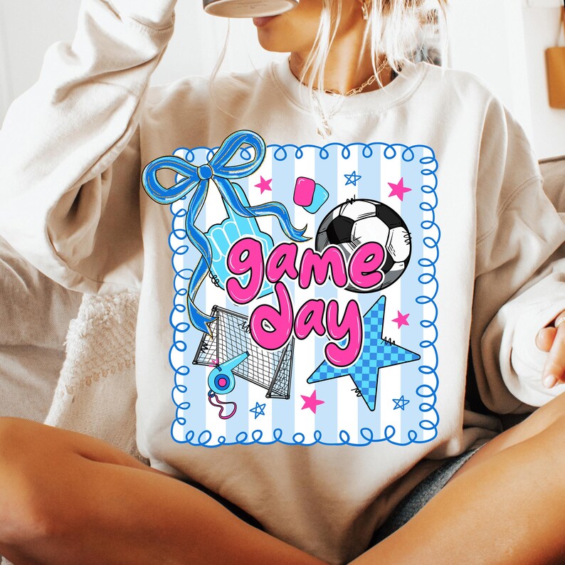 May include: Cream-colored sweatshirt with a colorful graphic. The graphic features a soccer ball, stars, a bow, and the words "Game Day" in pink. The background has blue and white stripes.