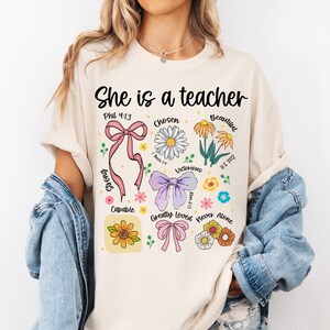 Op de afbeelding: Cr&egrave;mekleurig T-shirt met de tekst "She is a teacher" en inspirerende woorden en bloemenafbeeldingen. Het ontwerp bevat strikken, madeliefjes en andere bloemen, met zinnen als "Chosen" en "Beautiful".