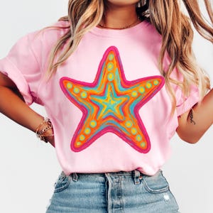 May include: Pink t-shirt featuring a large, colorful starfish design. The starfish has a pink outline, orange, yellow, and blue interior, and yellow dot accents. The shirt is made of a soft material and has rolled-up sleeves.
