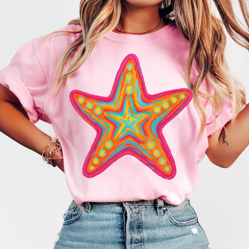 May include: Pink t-shirt featuring a large, colorful starfish design. The starfish has a pink outline, orange, yellow, and blue interior, and yellow dot accents. The shirt is made of a soft material and has rolled-up sleeves.