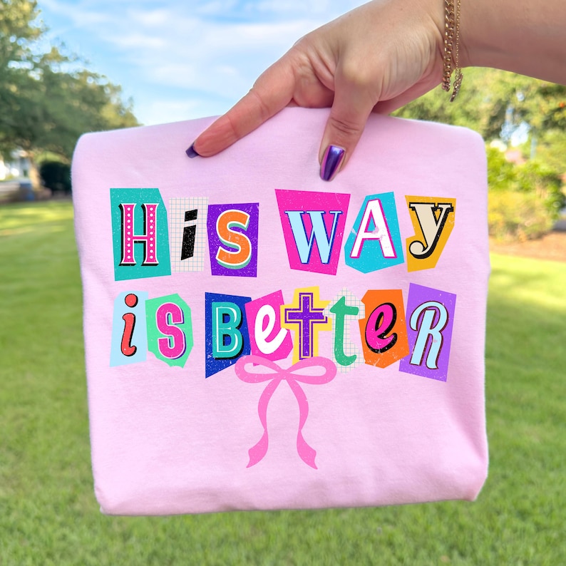 May include: A light pink sweatshirt with the text "His WAY is BETTER" in colorful, cut-out style letters. A pink bow is at the bottom of the design. The sweatshirt is held up by a hand.