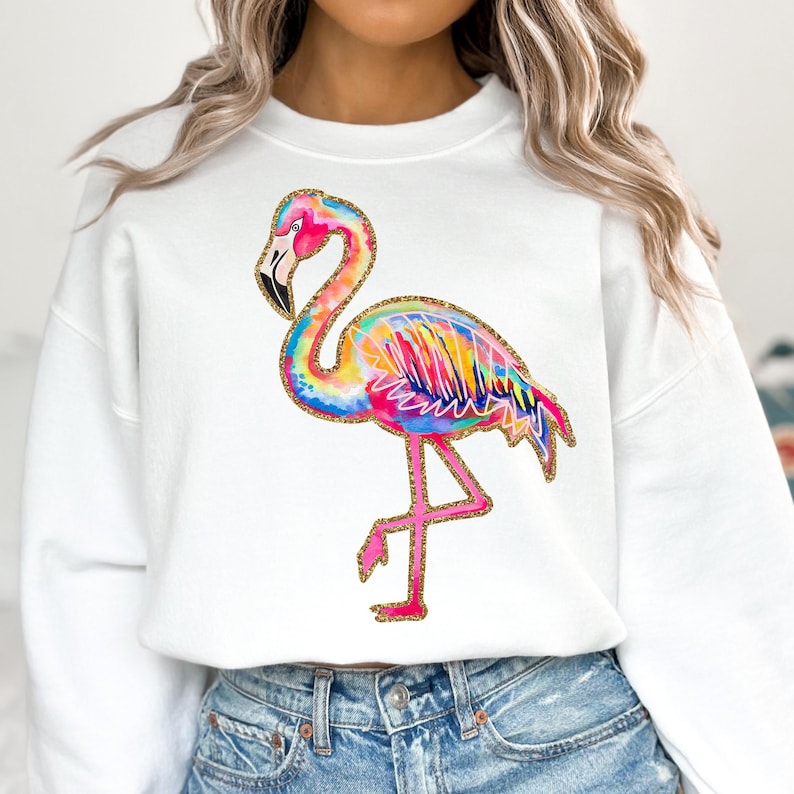 May include: White sweatshirt featuring a colorful flamingo design. The flamingo is depicted in vibrant shades of pink, blue, yellow, and orange, with gold glitter accents. The bird is standing on one leg, creating a playful and eye-catching design.