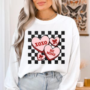 May include: White sweatshirt with a checkered pattern and pink hearts with red glitter. The hearts have the text "XOXO" and "BE MINE" on them.