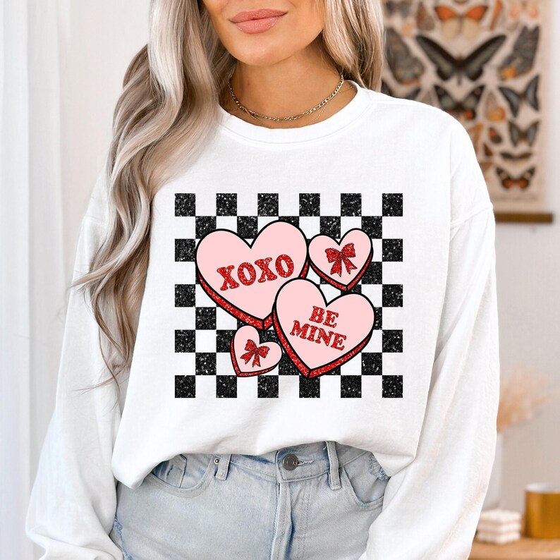 May include: White sweatshirt with a checkered pattern and pink hearts with red glitter. The hearts have the text "XOXO" and "BE MINE" on them.