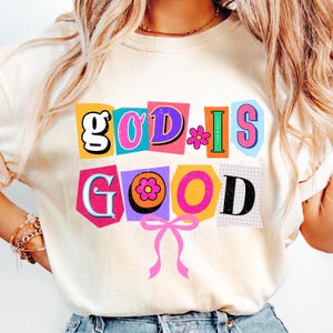 May include: Cream-colored sweatshirt with the phrase "GOD IS GOOD" in a colorful, cut-out style. The design includes flowers and a pink bow, creating a cheerful and positive aesthetic.