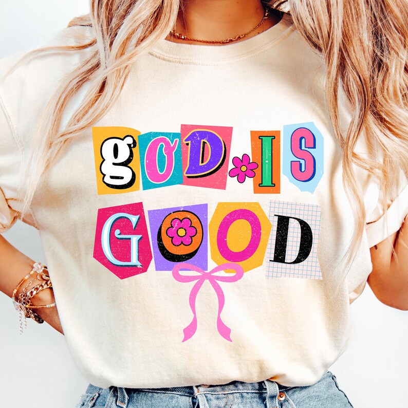 May include: Cream-colored sweatshirt with the phrase "GOD IS GOOD" in a colorful, cut-out style. The design includes flowers and a pink bow, creating a cheerful and positive aesthetic.