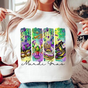 May include: White sweatshirt with a colorful Mardi Gras design featuring a mask, a crawfish, a king cake, and a jester's hat. The text "Mardi Gras" is printed below the design.