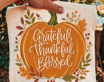 May include: A cream-colored t-shirt featuring a large orange pumpkin graphic with a green stem. The text Grateful Thankful Blessed is written in white cursive script across the pumpkin. The design is surrounded by autumn leaves and berries.