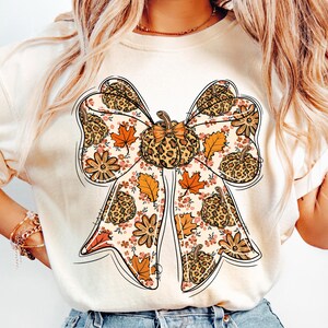 May include: Cream-colored t-shirt featuring a large bow design with a pumpkin and fall leaf theme. The bow has a leopard print pattern, and the design includes orange, brown, and pink floral accents. The overall style is autumnal.