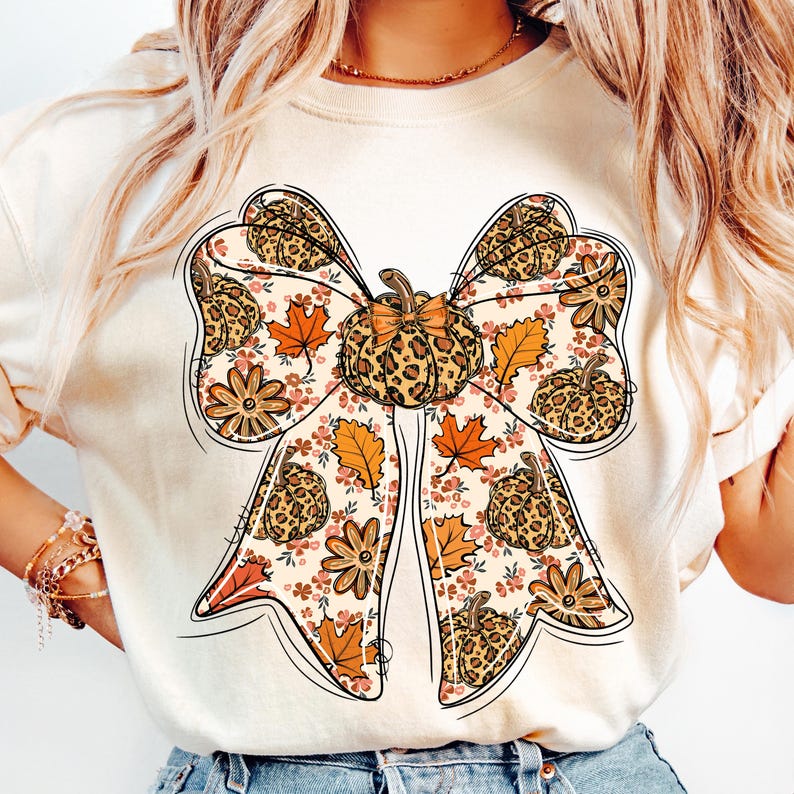 May include: Cream-colored t-shirt featuring a large bow design with a pumpkin and fall leaf theme. The bow has a leopard print pattern, and the design includes orange, brown, and pink floral accents. The overall style is autumnal.